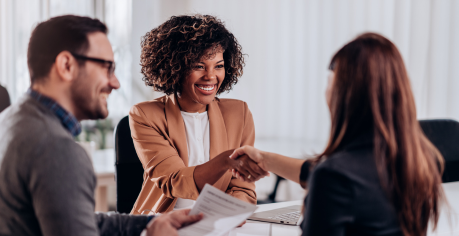 Two women shaking hands in a business meeting while male colleague smiles on the left.