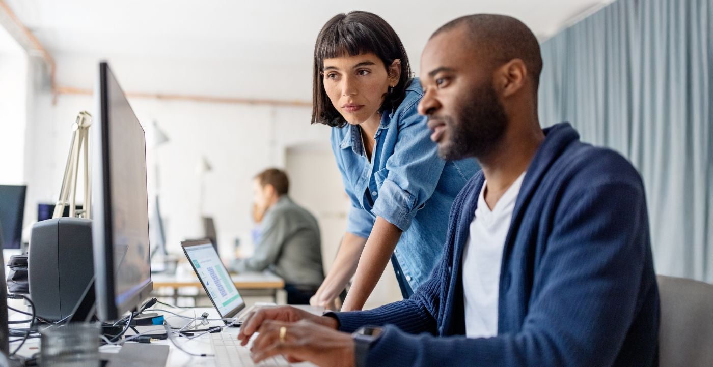 Two people look at a computer screen in an office setting. The person in the foreground is typing while the other looks on. Another person is visible in the background.