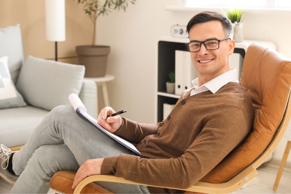 A man wearing glasses and a brown cardigan sits in a leather chair, holding a clipboard and pen.
