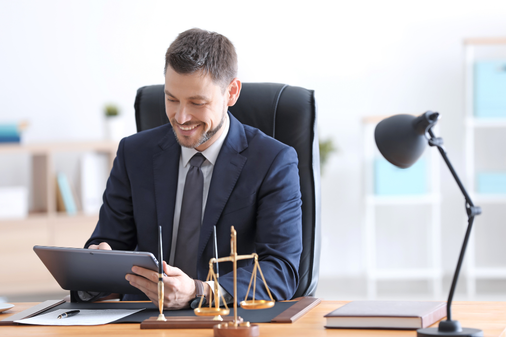 A man in a suit sits at a desk, smiling and looking at a tablet. On the desk are a pen, a scale of justice, and a lamp.