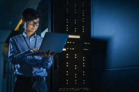 IT professional checking a laptop in a data center with blue lighting and server towers.