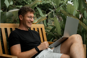 Smiling man sitting on a wooden bench outdoors, working on a laptop surrounded by green tropical plants