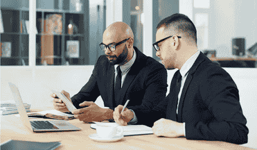 Two professionals in suits collaborating at a table with a laptop and documents.