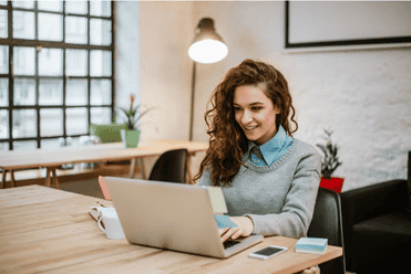 A smiling woman with curly hair working on her laptop at a shared workspace.