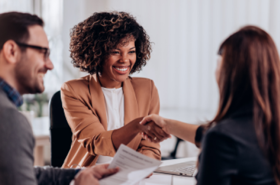 Two women shaking hands in a business meeting while male colleague smiles on the left.