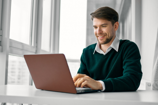 A man wearing a green sweater and white shirt smiles as he types on a maroon laptop at a white desk in front of a window.