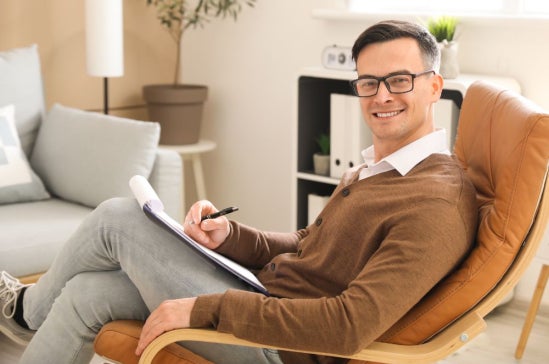 A man wearing glasses and a brown cardigan sits in a leather chair, holding a clipboard and pen.