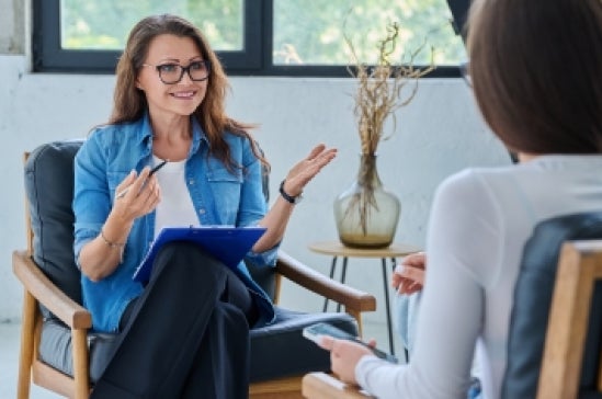 A woman in a denim shirt and glasses holds a clipboard and pen while speaking to another person in a therapy session.