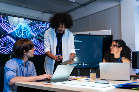Three people are gathered around a table in a dimly lit room, with a large screen displaying a blue, circuit-board-like brain graphic behind them. 