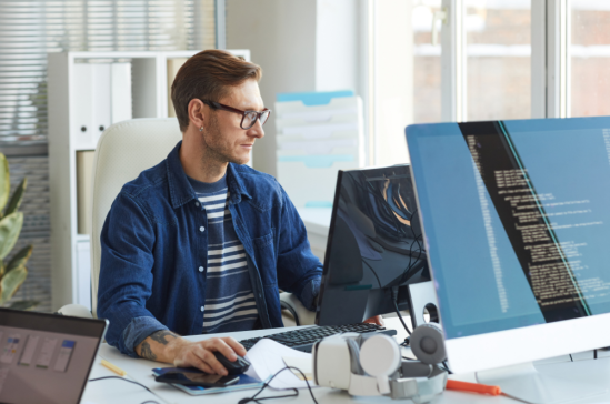 A person wearing glasses and a blue shirt sits at a desk, working on a computer with multiple monitors displaying code.