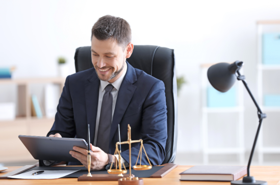 A man in a suit sits at a desk, smiling and looking at a tablet. On the desk are a pen, a scale of justice, and a lamp.