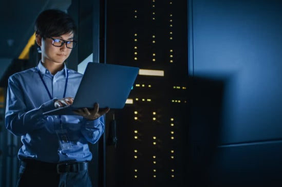 IT professional checking a laptop in a data center with blue lighting and server towers.