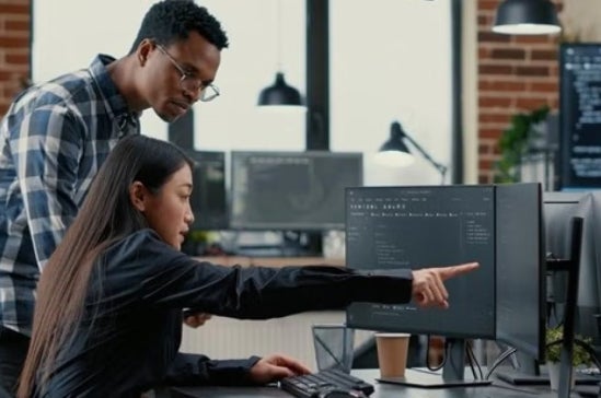 Two people looking intently at a computer screen in an office setting, the woman sitting and pointing while man stands looking at the same screen.