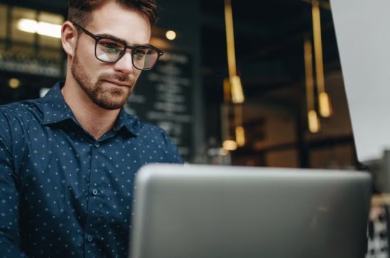 Young man with glasses works on a laptop in a modern café or workspace, representing starting a career in finance or entry-level job searches.