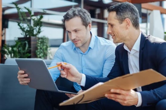 Two men in business attire sit together in a modern office lounge, discussing and reviewing information on a laptop and a folder.