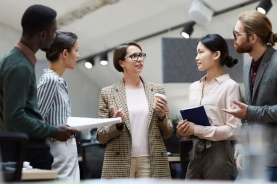 Group of five diverse professionals talking together in a modern office setting, suggesting workplace collaboration and communication.