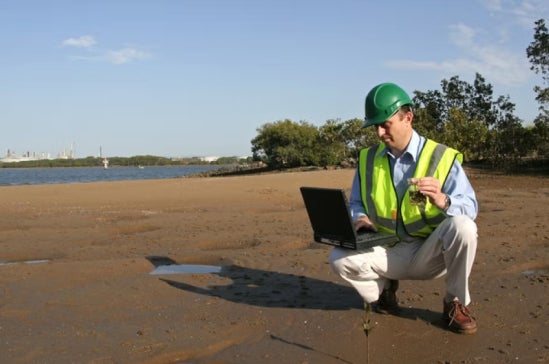 An environmental scientist stood on mudflats analysing data on his laptop