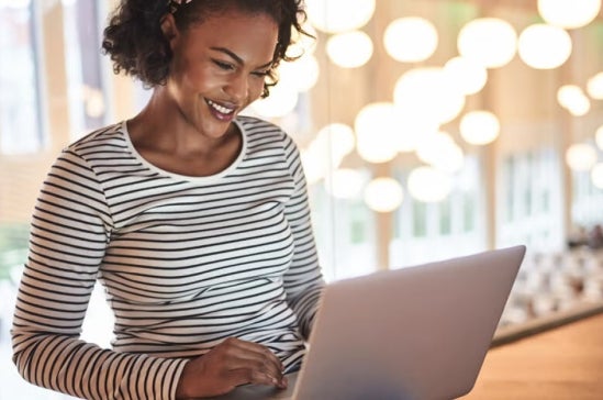 Woman in a striped shirt using a laptop in a brightly lit room, suggesting online research or studying financial aid options.