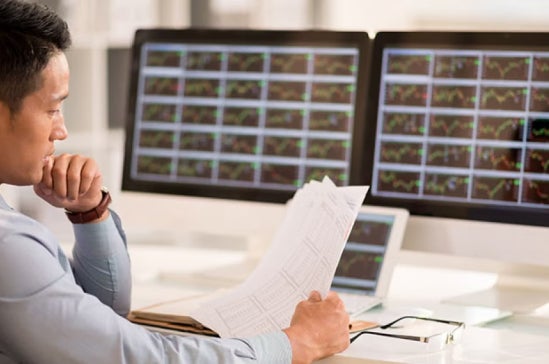 Man in business attire analyzes market data across multiple monitors, holding data sheets, illustrating the rise of marketing analytics in finance.