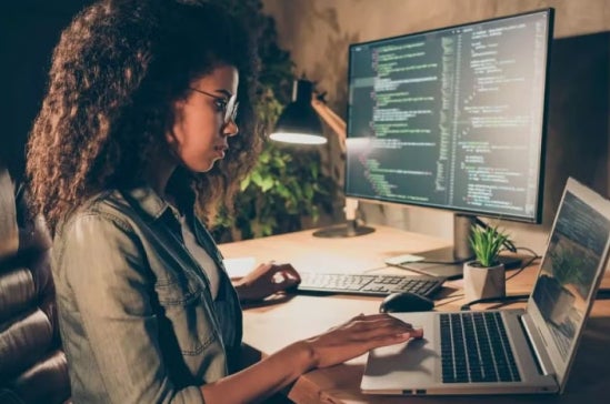 Woman with curly hair works at a desk with a laptop and large monitor displaying code, highlighting female representation in cybersecurity and tech.