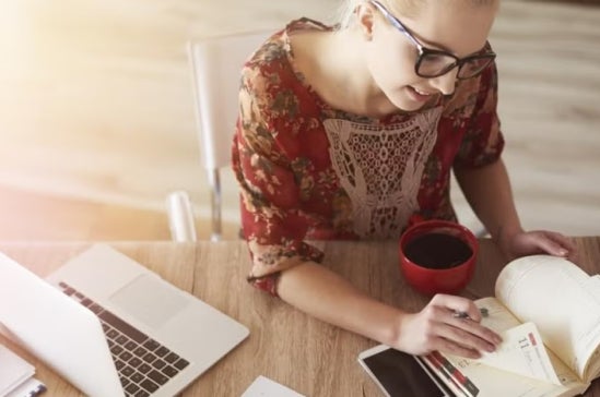 Woman in a floral dress sits at a desk with a planner and coffee, using a laptop, suggesting balancing work tasks and study in a home or casual workspace