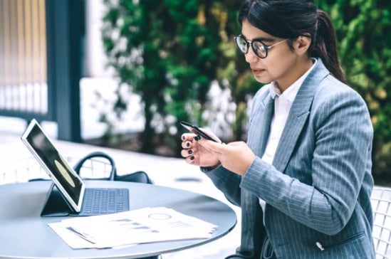 Businesswoman in a pinstripe suit sits outside at a table with a tablet, reviewing financial documents and typing on her phone.