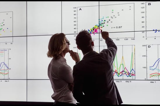 Two people analyze colorful data charts and graphs displayed on a large screen.