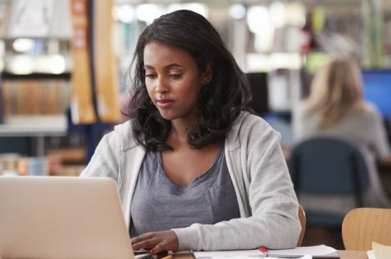 Woman working on a laptop in a public library, focused on her studies, suggesting career advancement or preparation for a finance manager role.