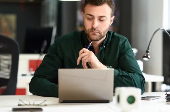Man in a green shirt sits in a modern office at a desk with a laptop, holding a pen thoughtfully, suggesting focus on analytical work or career planning.