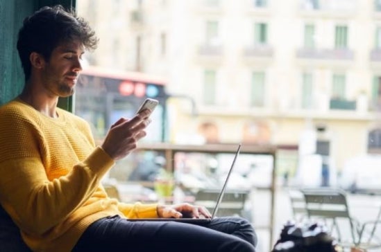 Young man in a yellow sweater sits by a window with a laptop, looking at his phone, suggesting remote work or digital job search in a city setting.