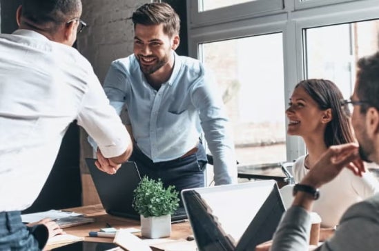 Group of professionals in a meeting room, one person stands and shakes hands with a colleague, while others smile and interact around laptops, suggesting leadership and teamwork