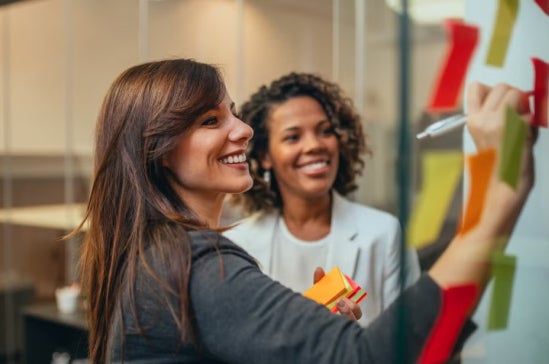 Two women smiling and collaborating while writing ideas on colorful sticky notes.