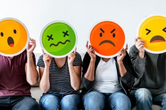 Four people sit side by side holding large emoji faces in front of their heads, each emoji showing a different emotion: surprise, sickness, anger, and sadness.