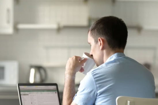 Man in a light blue shirt sits at a kitchen table with a laptop, drinking from a cup, suggesting remote learning or online study in a home environment.