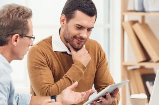 Two men in discussion at a desk, one holding a tablet and the other gesturing, with bookshelves and a laptop in the background, illustrating academic or educational consultation.