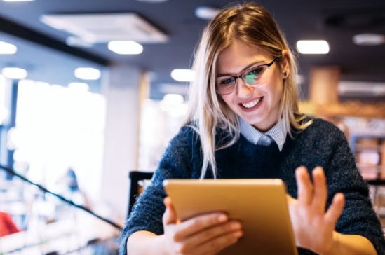 Woman with long blonde hair uses a tablet in a modern cafe or workspace, illustrating participation and advancement of women in the finance sector.