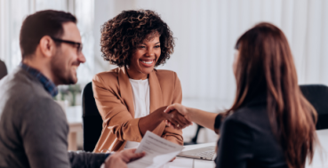 Two women shaking hands in a business meeting while male colleague smiles on the left.