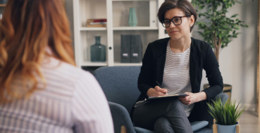A therapist sits in a chair, holding a clipboard and pen, looking at a patient whose back is to the camera.