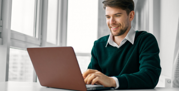A man wearing a green sweater and white shirt smiles as he types on a maroon laptop at a white desk in front of a window.