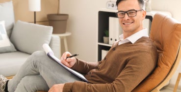 A man wearing glasses and a brown cardigan sits in a leather chair, holding a clipboard and pen.