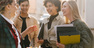 Four people are standing outside and talking. One person is on the phone. Three people are laughing. Two people are holding folders.