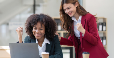 Two women in business attire celebrating success while looking at a laptop. One woman is seated and looking at the screen with her fist raised, while the other stands behind her, placing a hand on her shoulder with her own fist raised.
