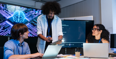Three people are gathered around a table in a dimly lit room, with a large screen displaying a blue, circuit-board-like brain graphic behind them. 