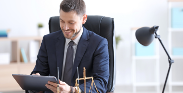 A man in a suit sits at a desk, smiling and looking at a tablet. On the desk are a pen, a scale of justice, and a lamp.