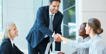 Business meeting in a bright office: a man in a suit stands and smiles while shaking hands with colleagues around a conference table with laptops and documents.