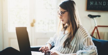 A woman sits in a brightly lit room working on a laptop, with a bicycle visible in the background.