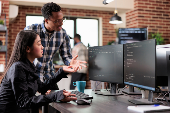 Two people looking intently at a computer screen in an office setting, the woman sitting and pointing while man stands looking at the same screen. 
