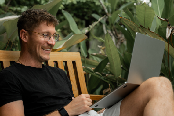 Smiling man sitting on a wooden bench outdoors, working on a laptop surrounded by green tropical plants