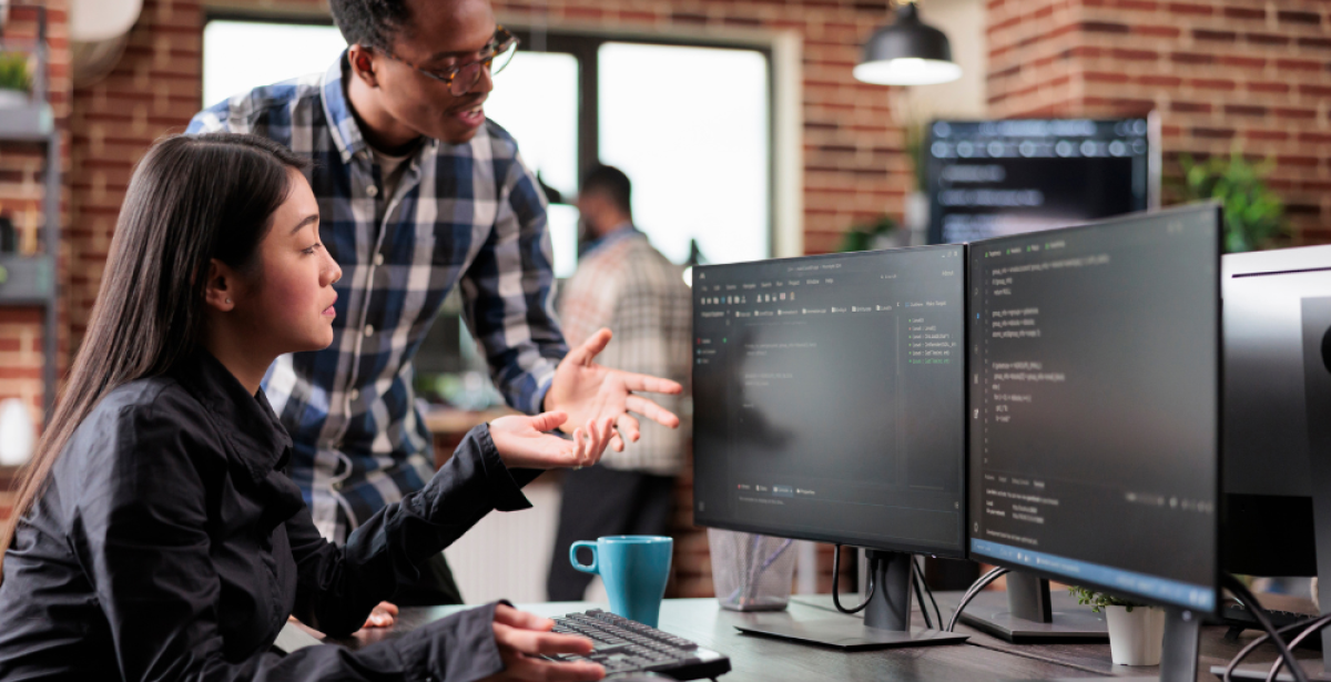 Two people looking intently at a computer screen in an office setting, the woman sitting and pointing while man stands looking at the same screen. 