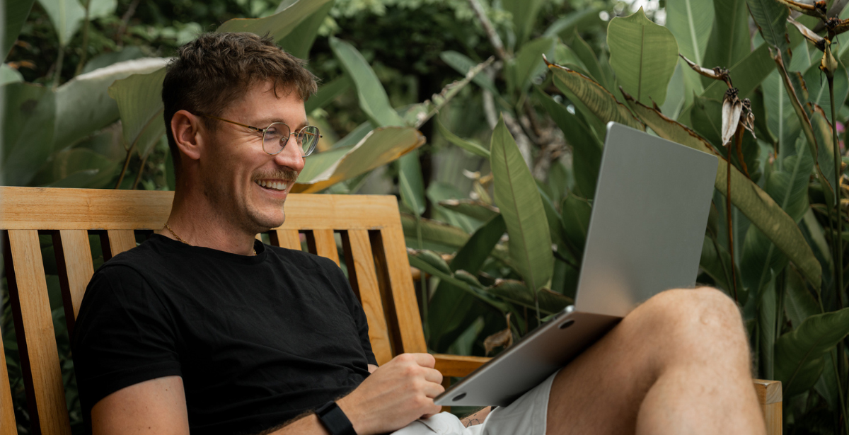 Smiling man sitting on a wooden bench outdoors, working on a laptop surrounded by green tropical plants